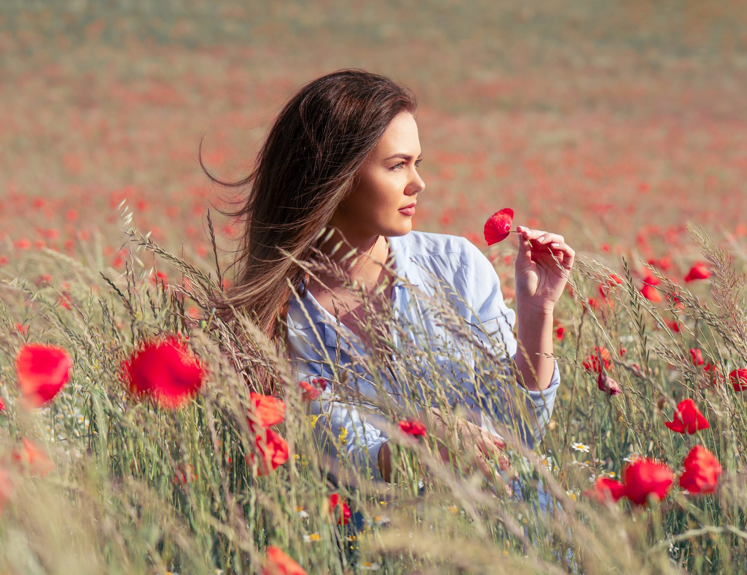 Eine junge Frau sitzt in einem Mohnfeld und riecht an einer Mohnblume.